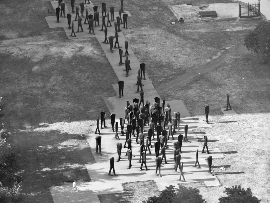 Aerial view of dozens of headless statues seemingly walking in every direction. A lone man walks through them.