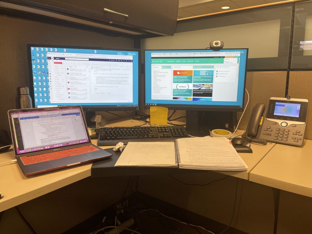 Several monitors display different work tasks inside an office cubicle